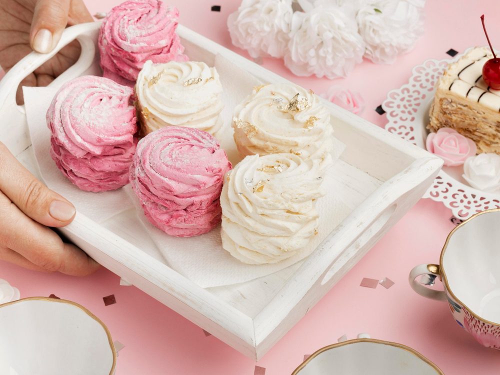 Merengues y torta servidos en bandeja blanca, con tazas y flores sobre fondo rosado